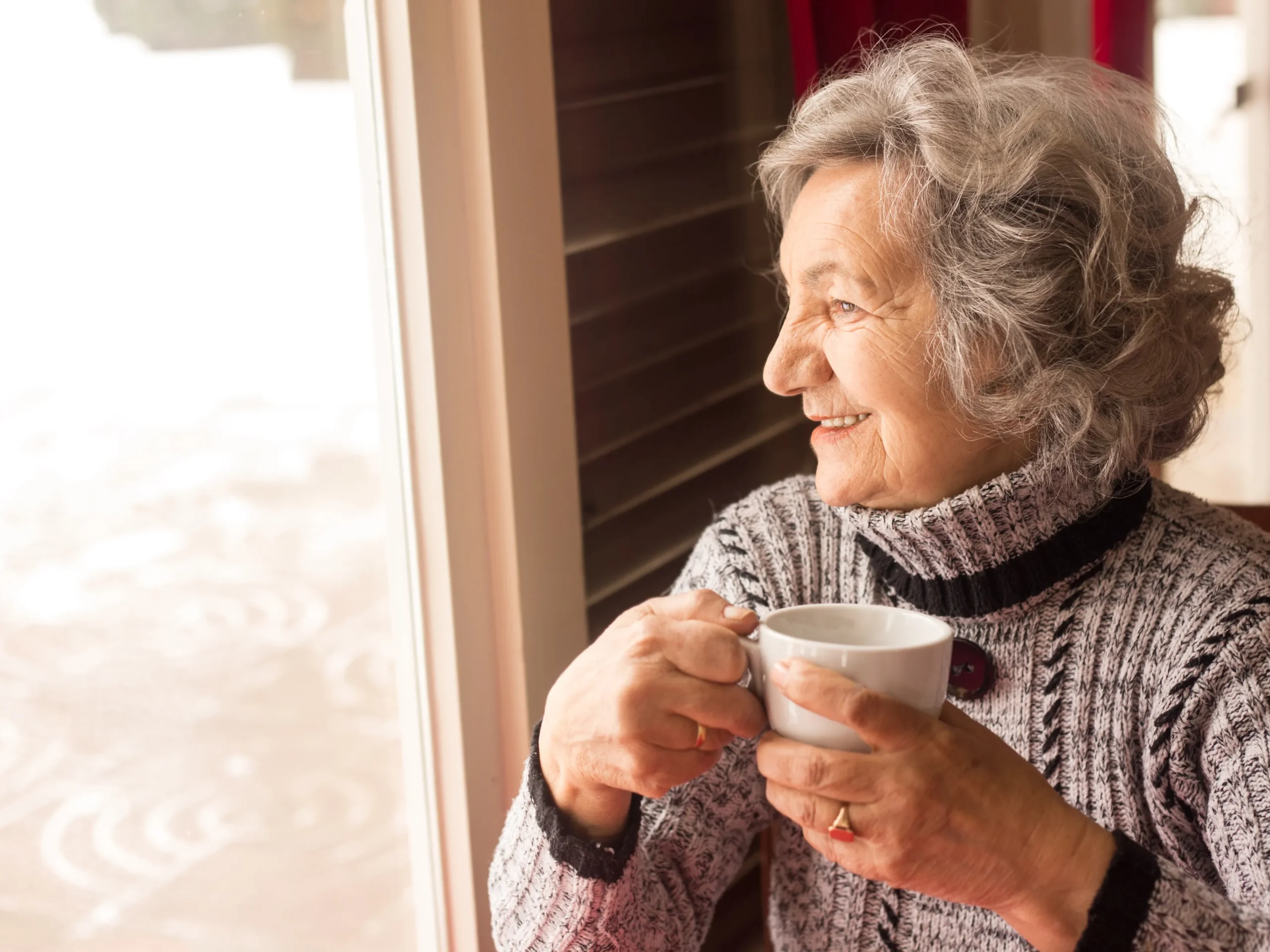 elderly woman enjoying hot drink while looking out the window in the winter